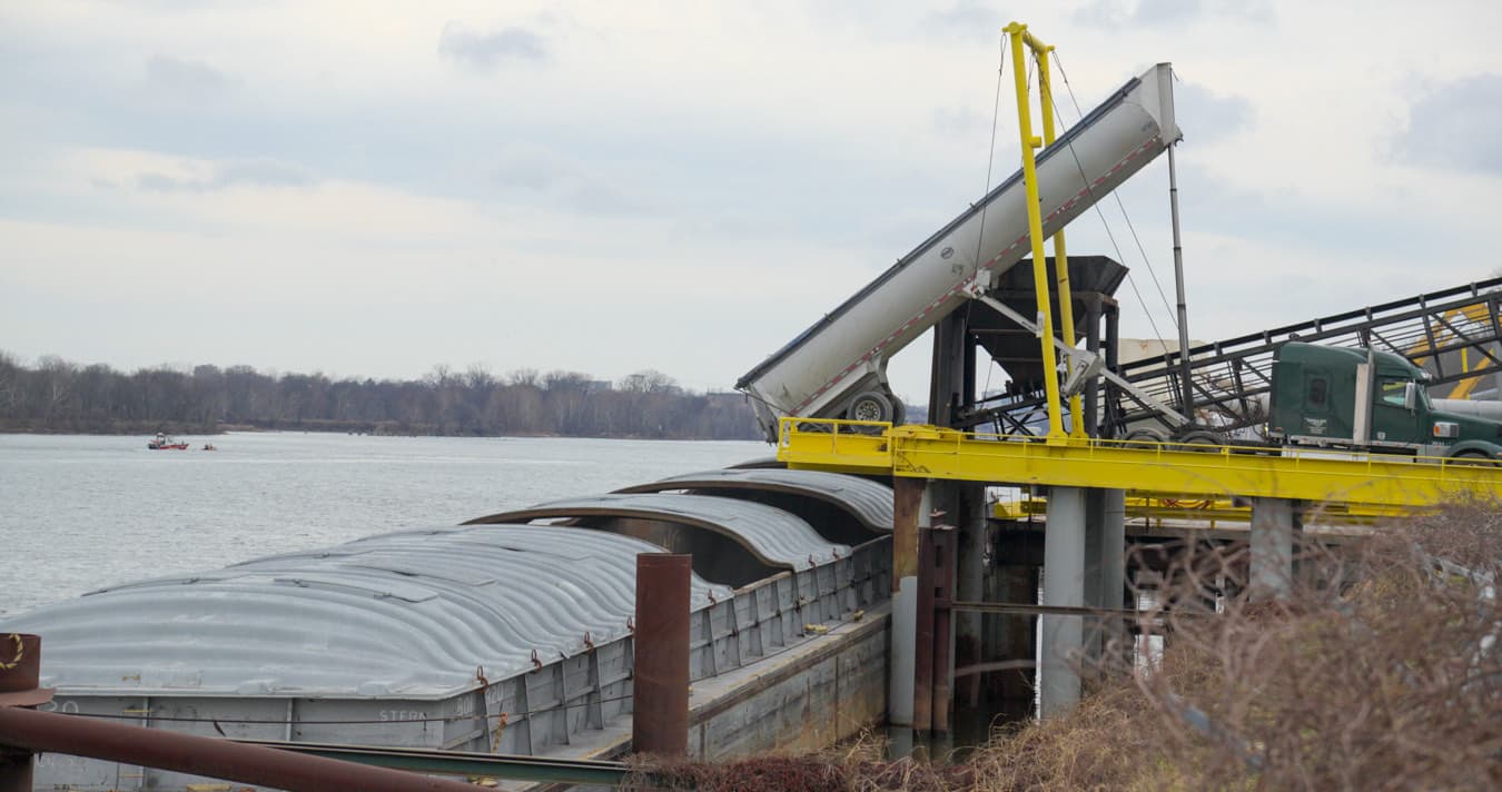 An end dump trailer discharging under a conveyor at a river terminal, with the load covered.