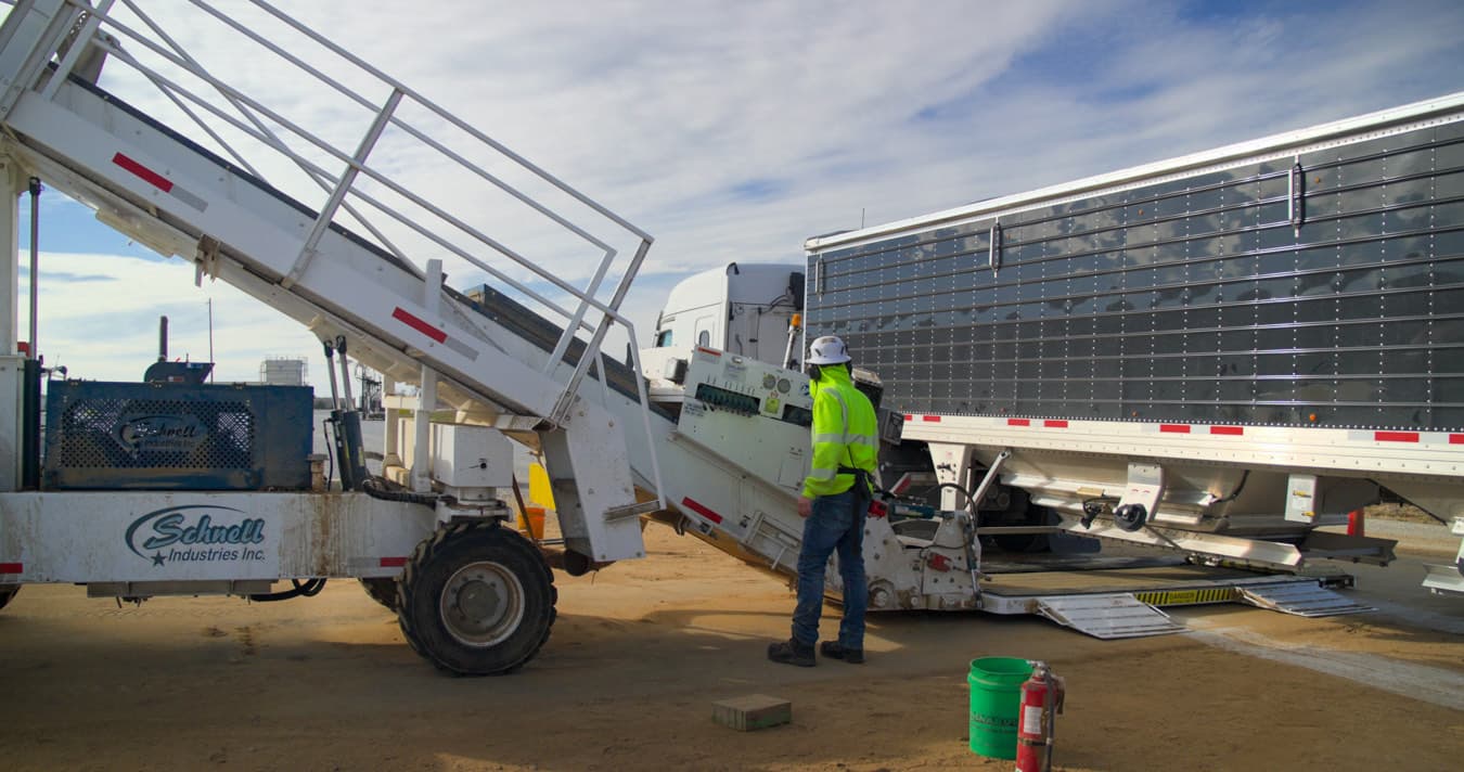 A hopper bottom trailer unloading grain into a receiving pit, with grain spilled across the ground and an operator standing alongside.