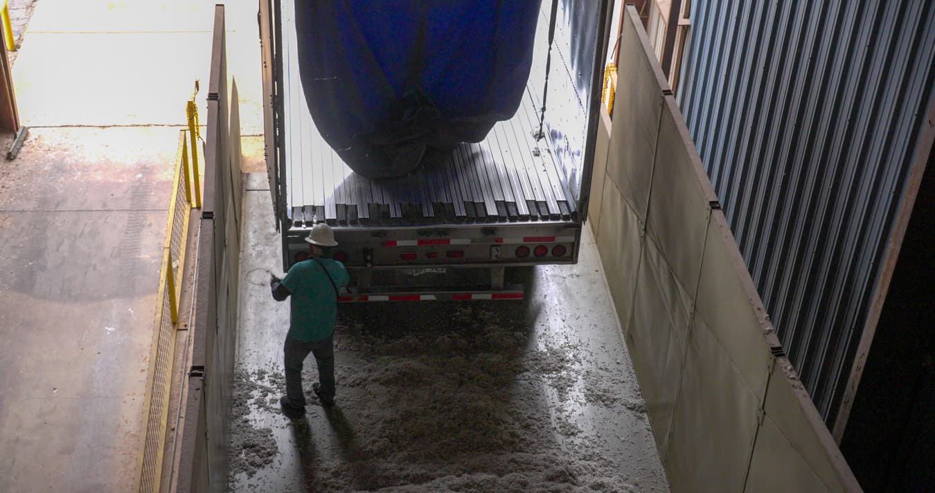 A walking floor trailer backed into a bay, discharging its load onto the floor while a worker clears behind it.