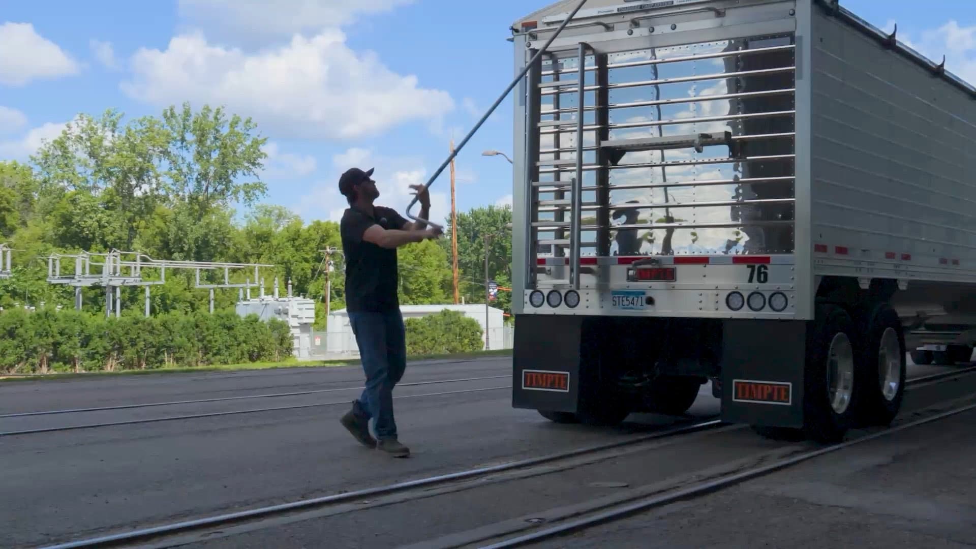 A driver pulling the tarp open across the top of a hopper bottom trailer on a summer afternoon.