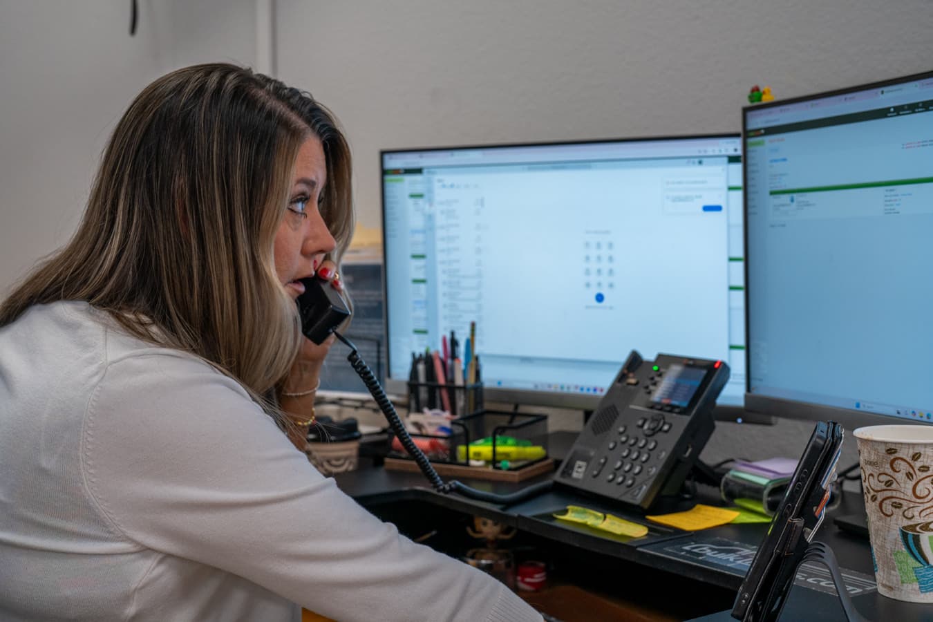 An account manager taking a call on a desk phone in front of two monitors.